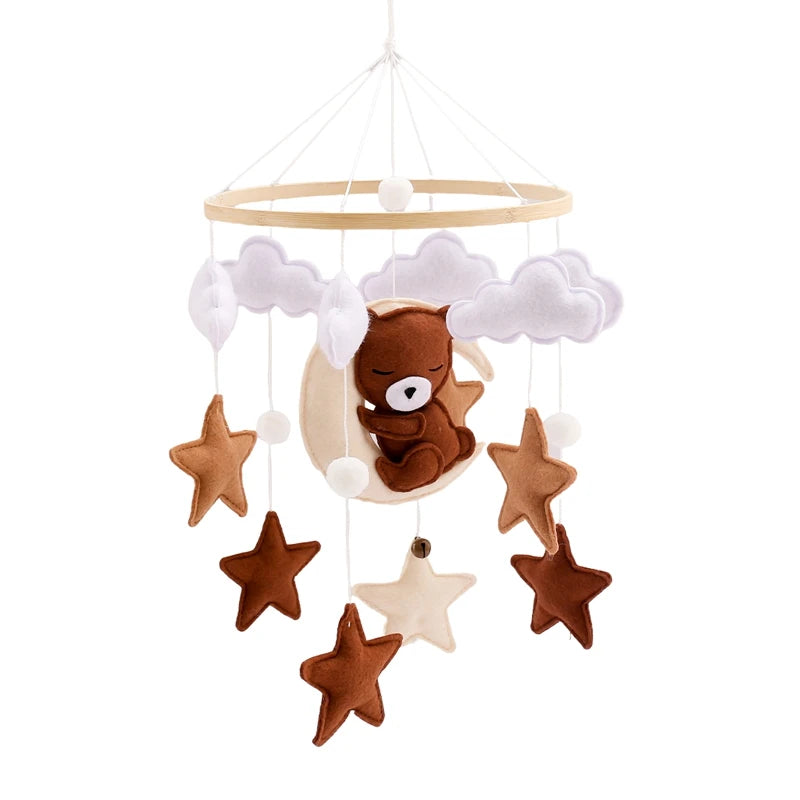 A whimsical baby mobile featuring a plush bear asleep on a crescent moon, surrounded by soft clouds and dangling stars in shades of brown and cream.