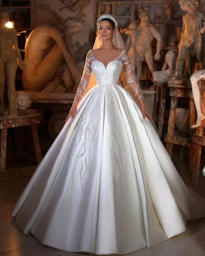 Bride in an elegant white gown with sheer lace sleeves and intricate embroidery, standing in an art studio surrounded by sculptures and dramatic lighting.