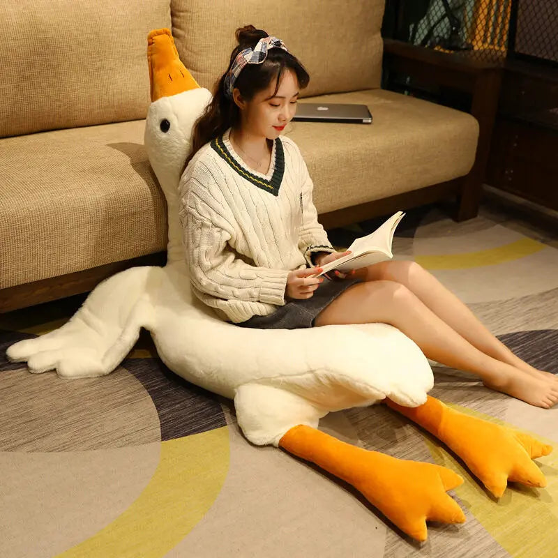 A woman sits on a plush, giant goose pillow while reading a book. She is wearing a knitted sweater and headband, seated on a carpeted floor in a cozy living room.