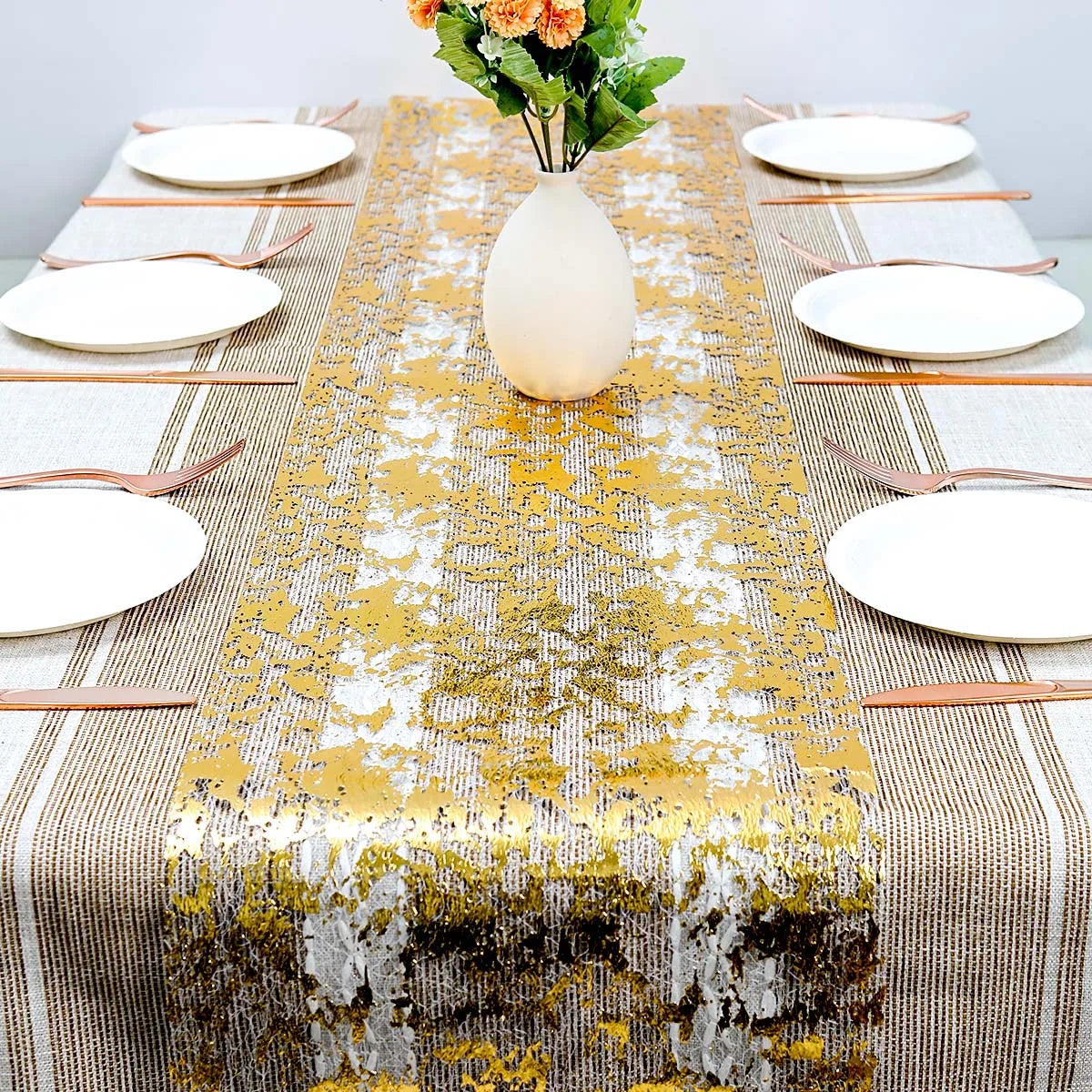 Elegant dining table set with a gold and white table runner, six white plates, and copper cutlery. A vase with orange flowers is placed centrally.