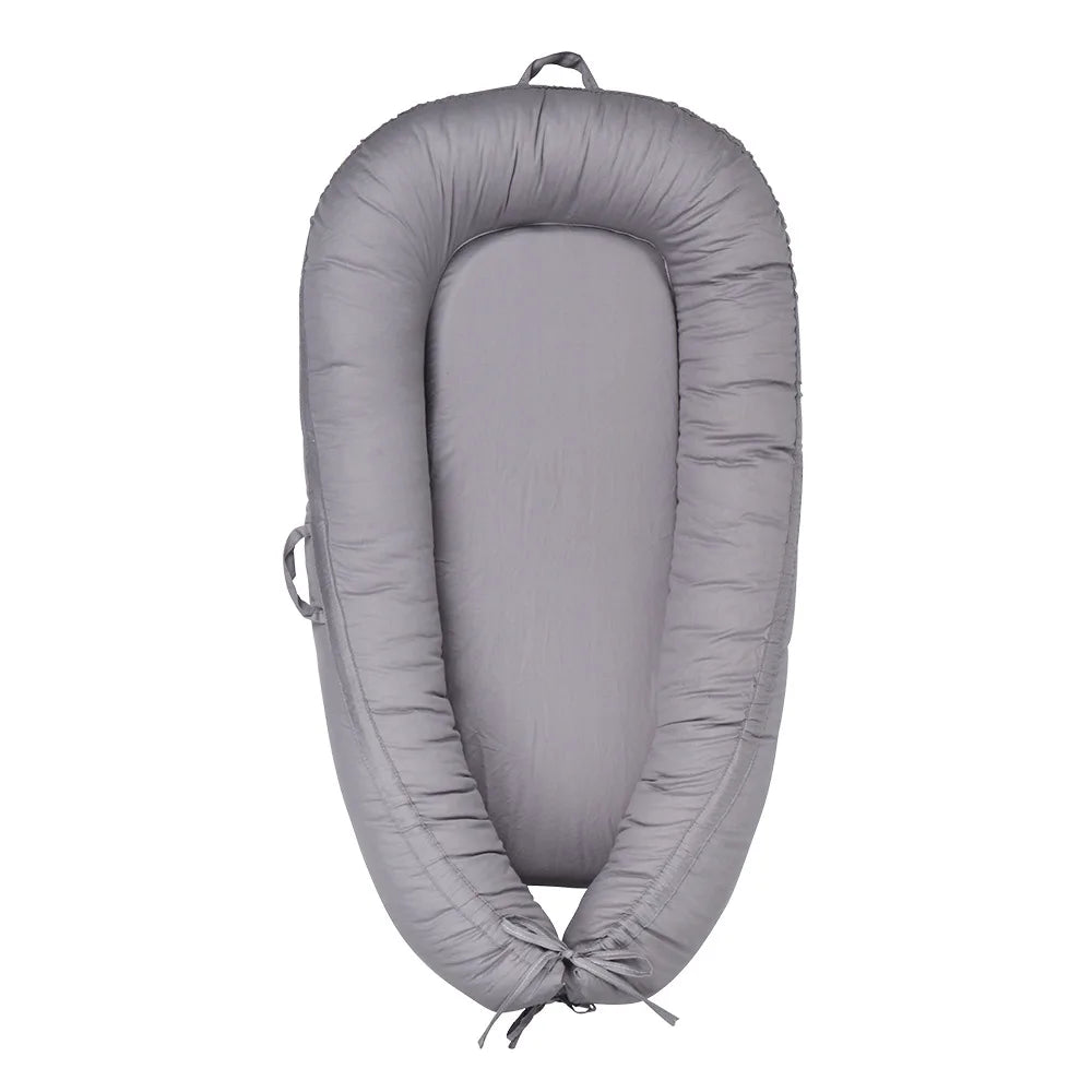 Gray baby lounger with a cushioned design and raised edges. Features handles on the sides and a tie at the bottom for adjustable snugness, providing a cozy space for infants.