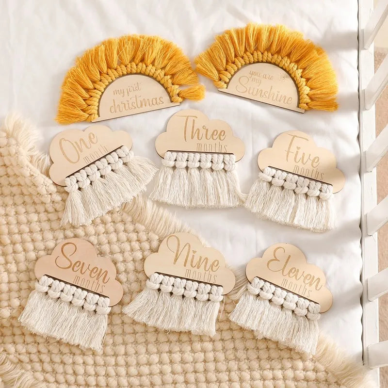 Wooden milestone plaques with white fringe and colorful yarn edging. Designs include month markers and phrases like "my first Christmas" on a textured beige blanket background.