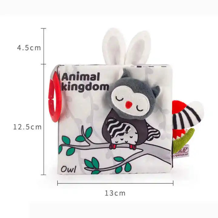 Soft fabric baby book titled "Animal Kingdom" featuring a cute owl design. It includes tactile elements like bunny ears and a red teething ring. Dimensions shown as 13cm by 12.5cm.