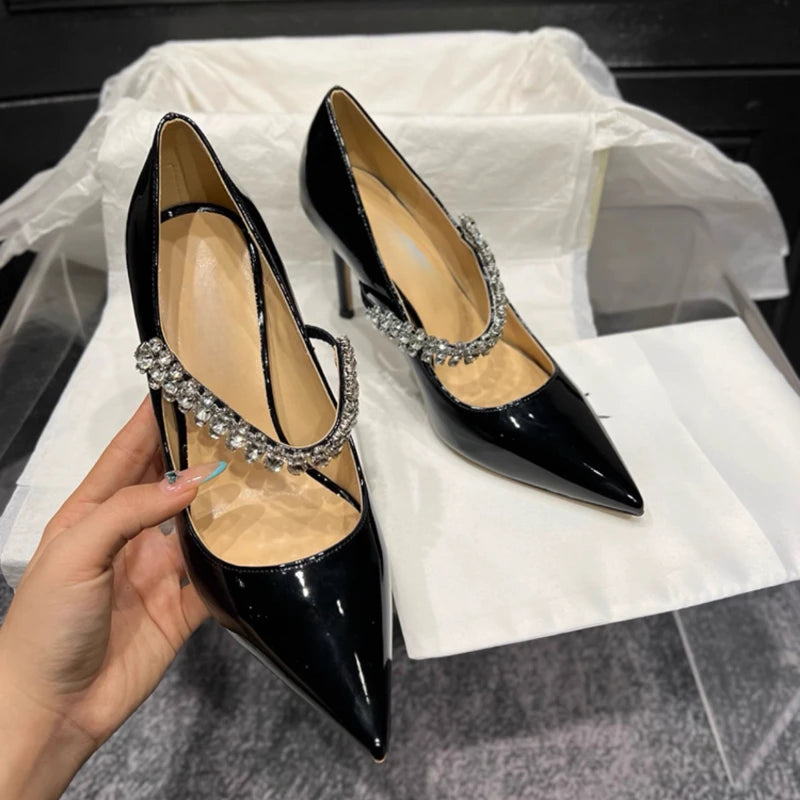 Black patent leather high heels with pointed toes and elegant crystal-embellished straps displayed on white paper. A hand gently holds one shoe in a stylish setting.