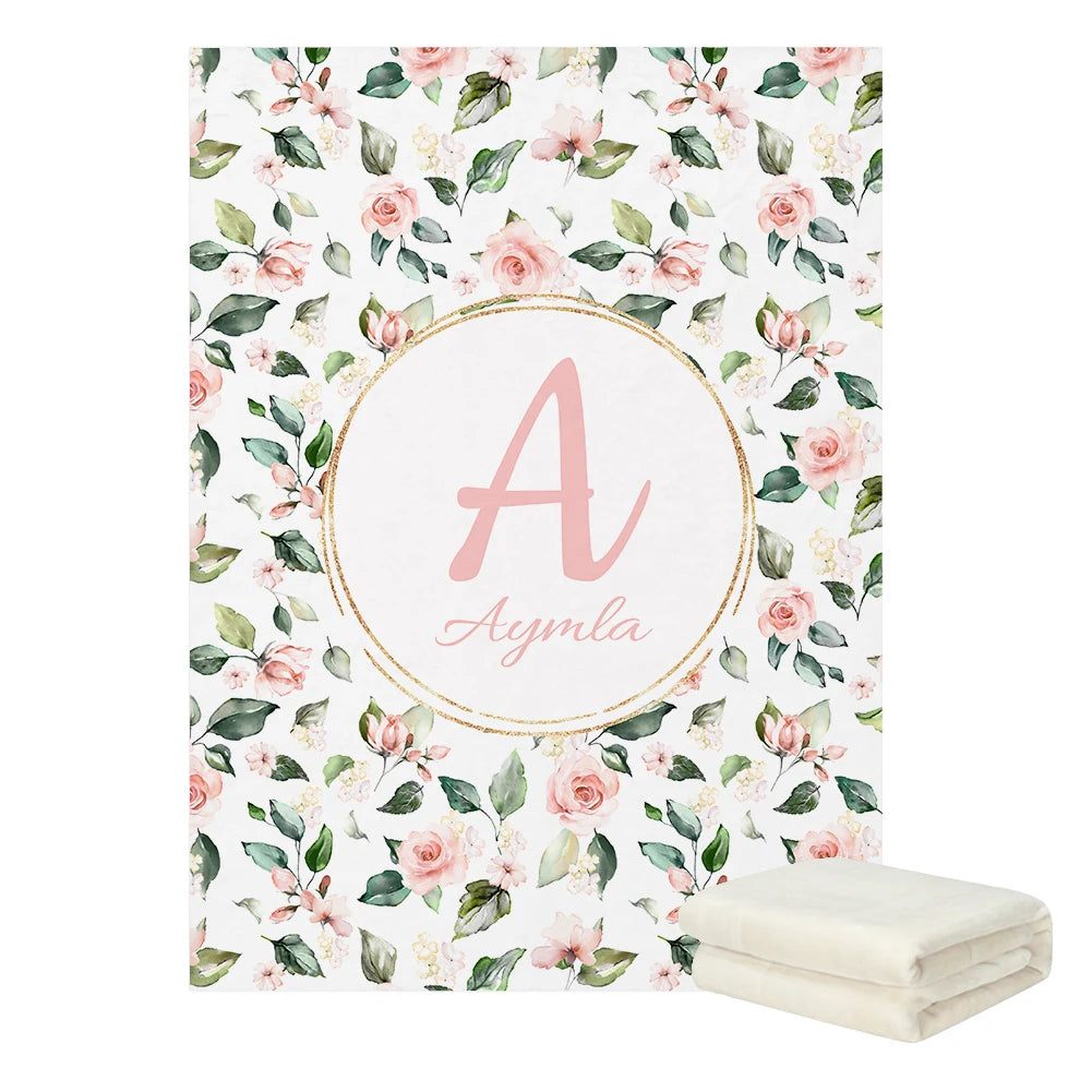 Floral blanket with pink roses and green leaves. Center features a large pink letter "A" and the name "Aymla" in script, encircled in gold. Folded white blanket on the side.