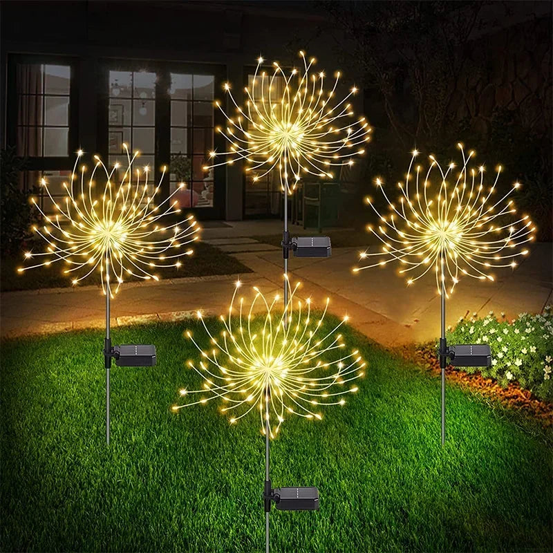 Four glowing dandelion-shaped solar lights illuminate a grassy yard at night, with a house in the background. The lights cast a warm, golden glow on the surrounding area.