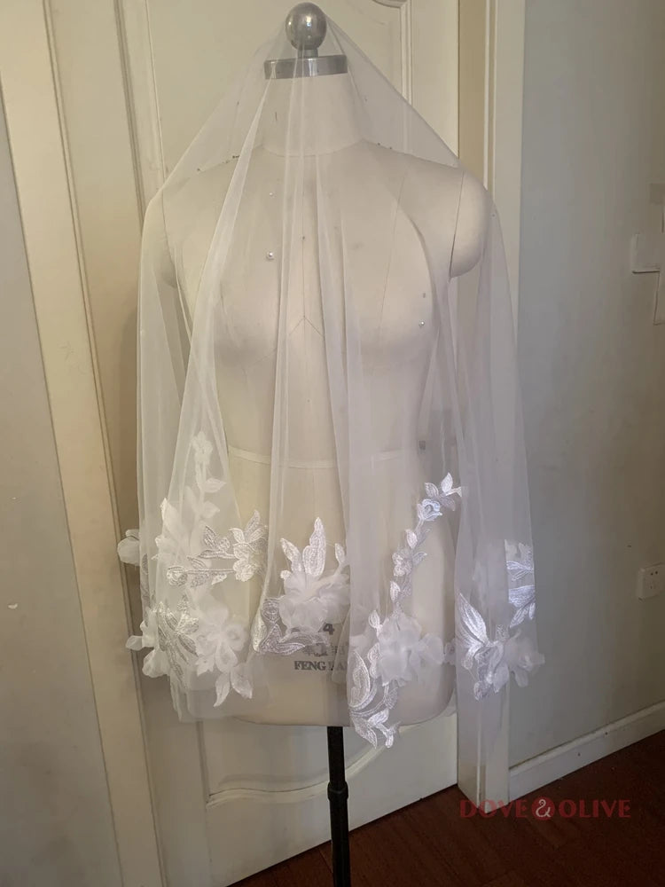 A mannequin displays a sheer bridal veil adorned with intricate white floral lace embroidery along the edges, set against a neutral background.
