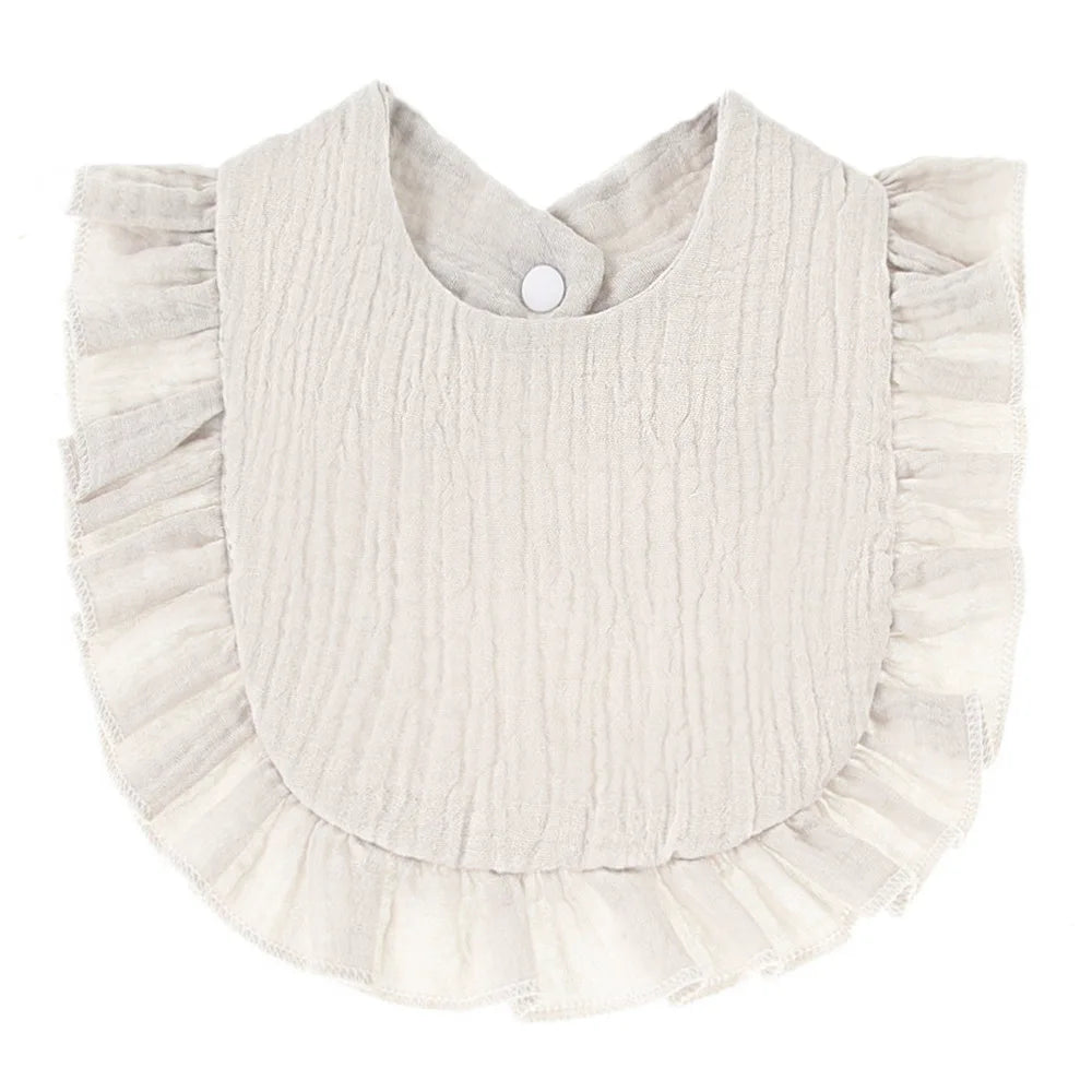 Cream-colored baby bib with a textured fabric and ruffled edges, featuring a round neckline and a snap button closure at the back.