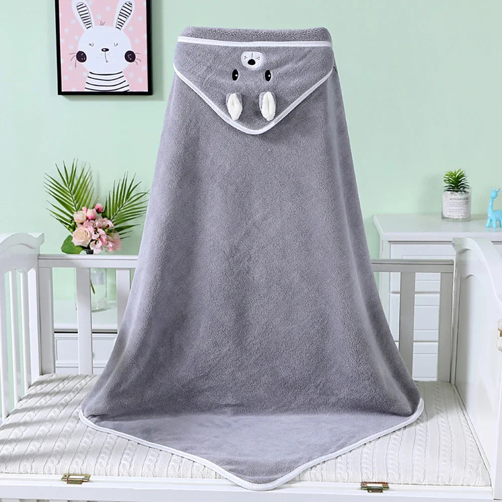 Gray hooded baby towel with animal face and ears on a crib. Background includes pastel green wall, framed rabbit artwork, pink flowers, and small plant.