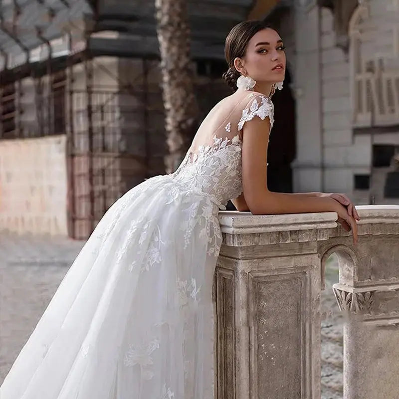 Bride in a lace wedding gown with cap sleeves leans on an ornate stone railing. The dress features intricate floral details with a flowing tulle skirt. Background shows historic architecture.