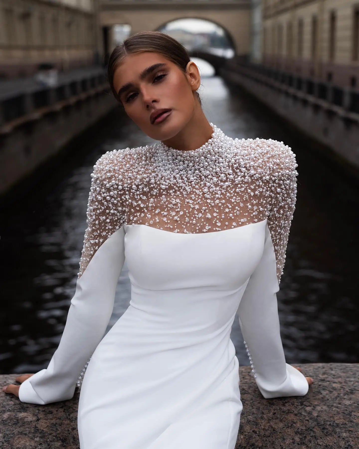 Woman in an elegant white gown with a pearl-studded sheer top, posed by a canal with arched bridges and historic buildings, exuding sophistication and grace.