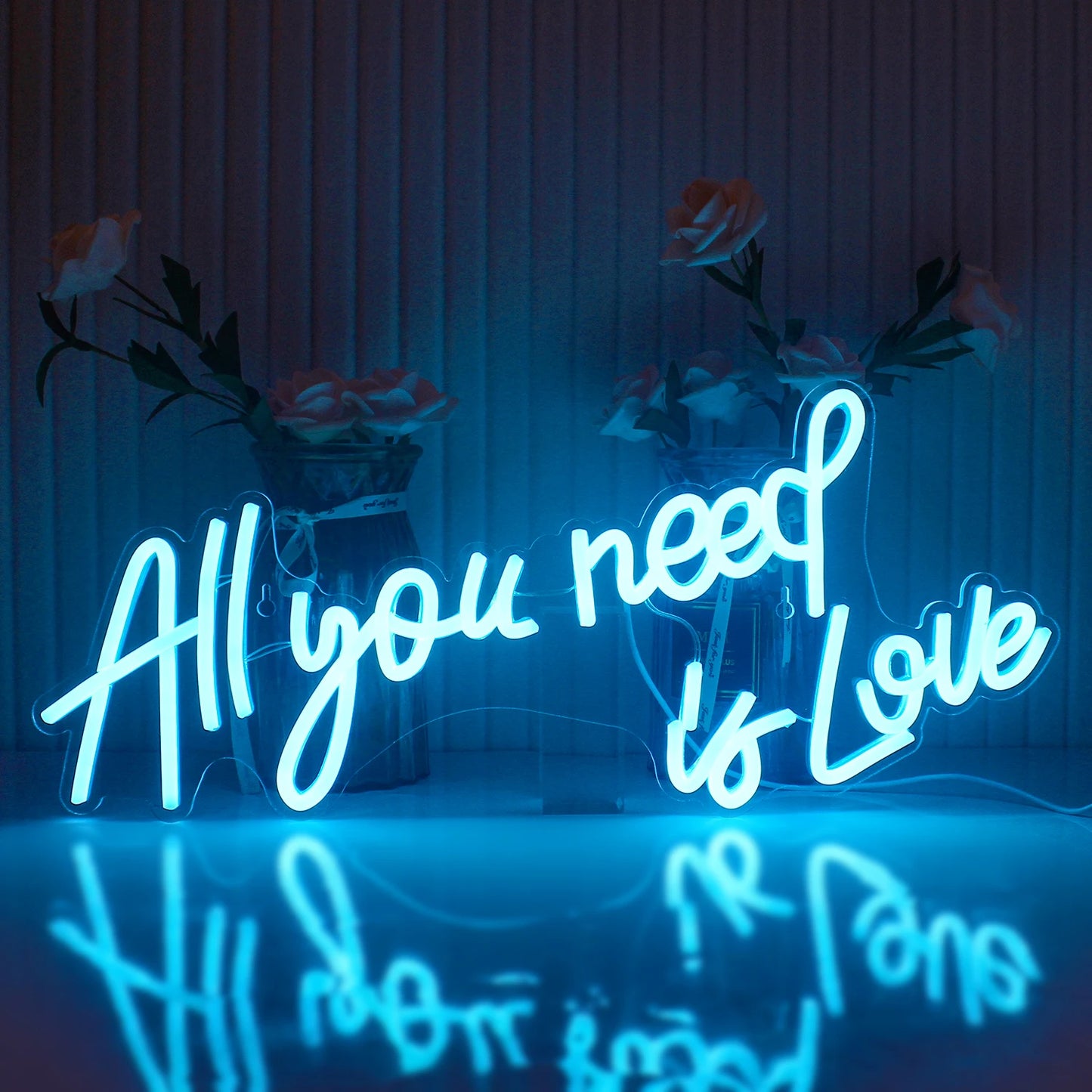 Blue neon sign reading "All you need is Love" with a reflection below, set against a background of vases with flowers.