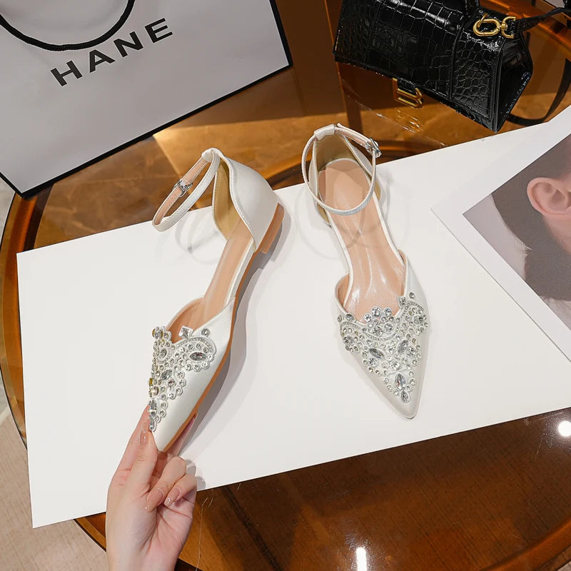 White pointed shoes with ankle straps adorned with sparkling gems are placed on a table. A hand holds one shoe. Nearby, a shopping bag and black purse can be seen.
