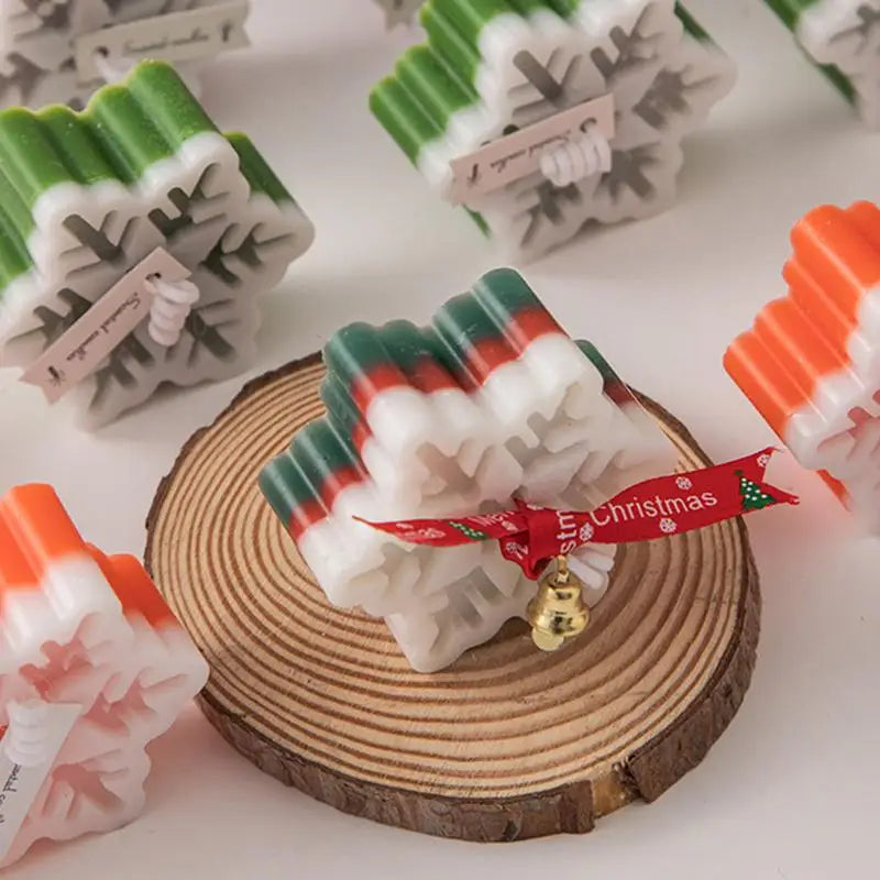 Festive snowflake-shaped soap bars in white, green, and red on a wooden slice. One soap has a red ribbon and a small gold bell attached, adding a Christmas touch.