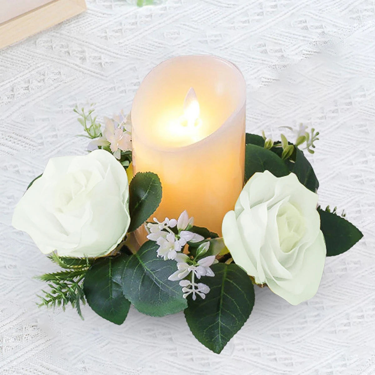 A glowing pillar candle is surrounded by white roses and green leaves, set on an intricately patterned white lace surface, creating an elegant and serene centerpiece.