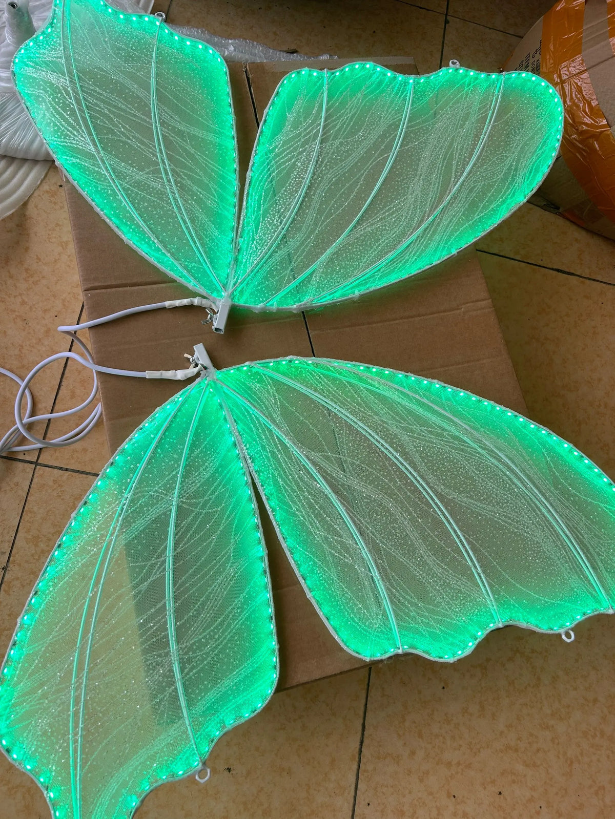 Two illuminated green translucent wings with glowing edges resembling fairy or insect wings, lying on a cardboard surface with a cardboard box nearby.
