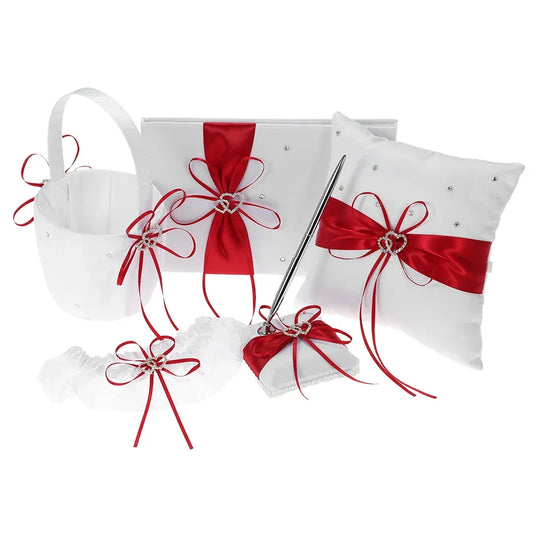 White wedding accessories with red ribbons and rhinestone heart accents, including a basket, guest book, ring pillow, garter, pen set, and flower girl basket.