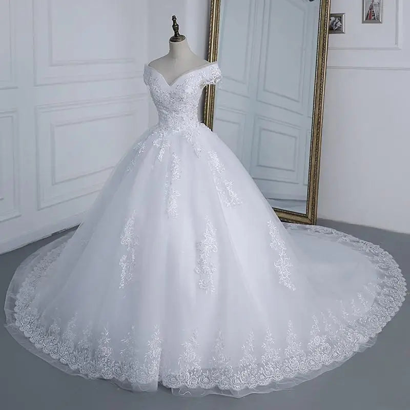 Elegant off-shoulder white wedding dress with lace details on a mannequin. The gown features a voluminous skirt and a long train, set in a room with a large mirror.
