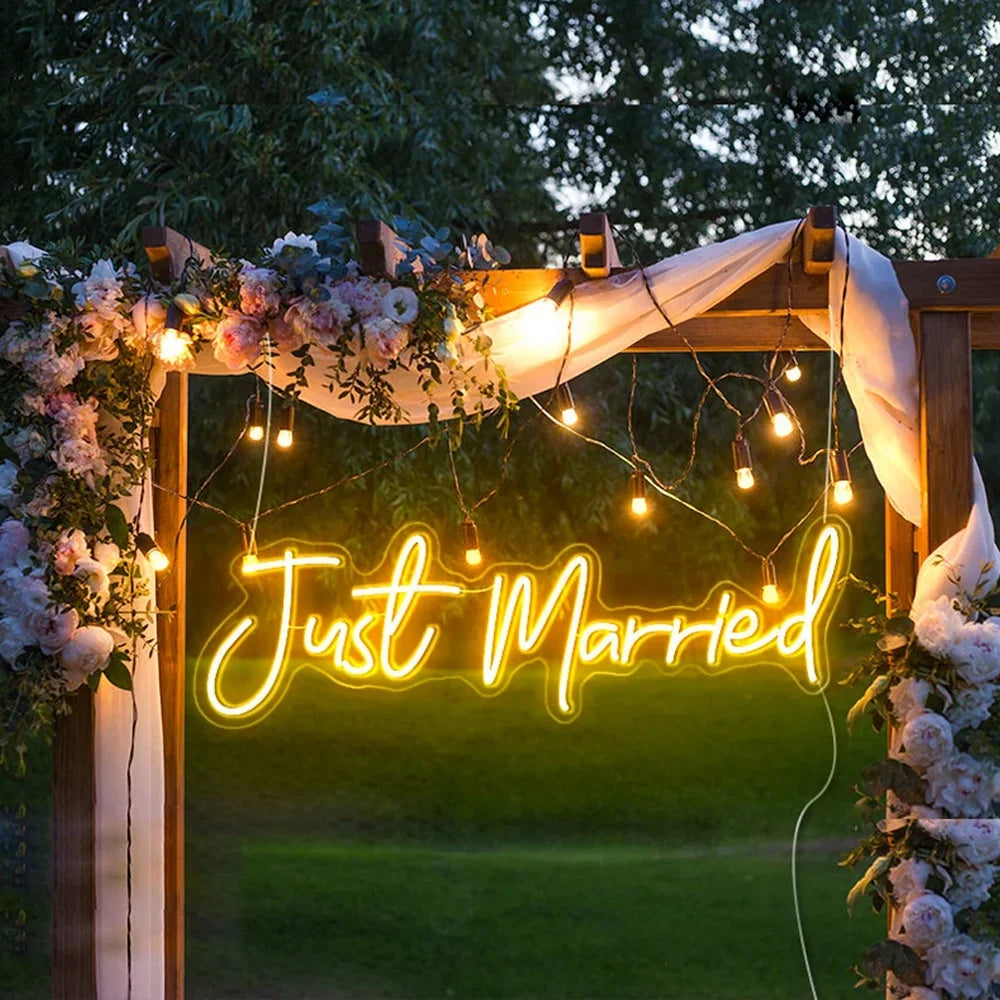 A wooden arch decorated with pink and white flowers, draped white fabric, and string lights frames a glowing "Just Married" neon sign against a greenery backdrop.