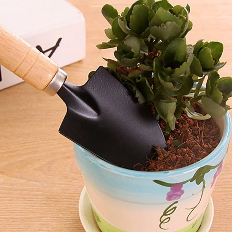 Small black garden trowel in pot with green plant and soil. Pot has blue and white colors with floral design, resting on light wooden surface.