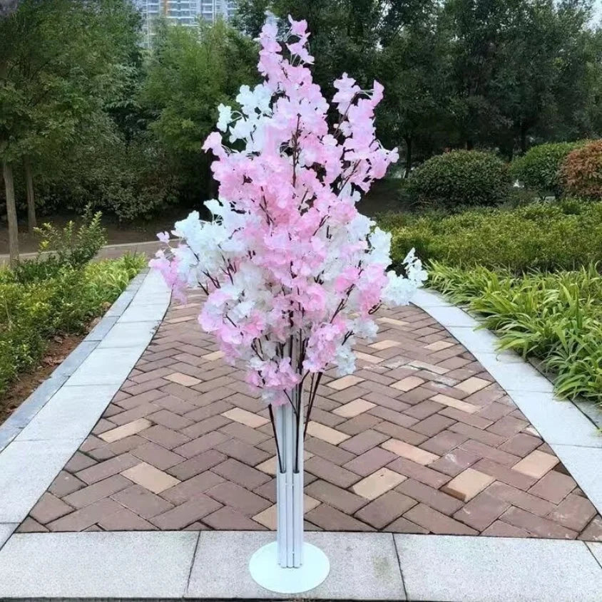 Artificial cherry blossom tree with pink and white flowers in a park, surrounded by lush greenery and a paved brick pathway.