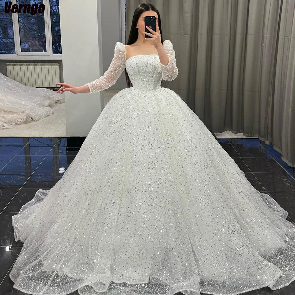 A woman in a fitting room takes a mirror selfie wearing a sparkling white ball gown with sheer, embellished long sleeves and a full skirt. The room has dark floors and curtains.