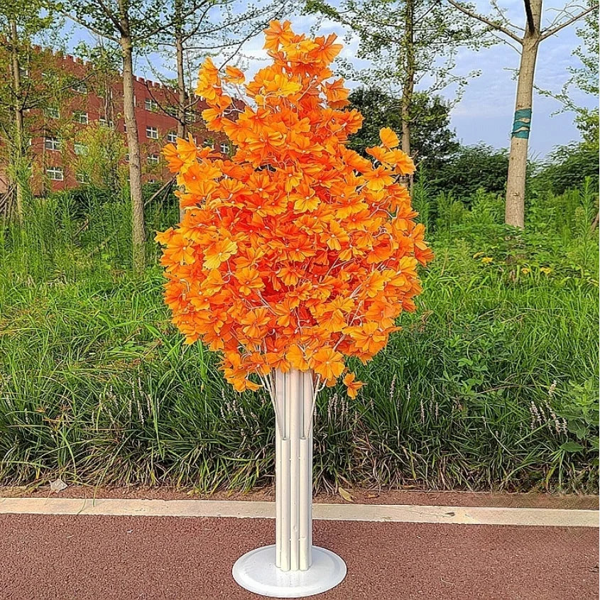 A vibrant artificial tree with bright orange flowers on a white stand, set against a backdrop of green foliage and a red brick building in a park-like setting.