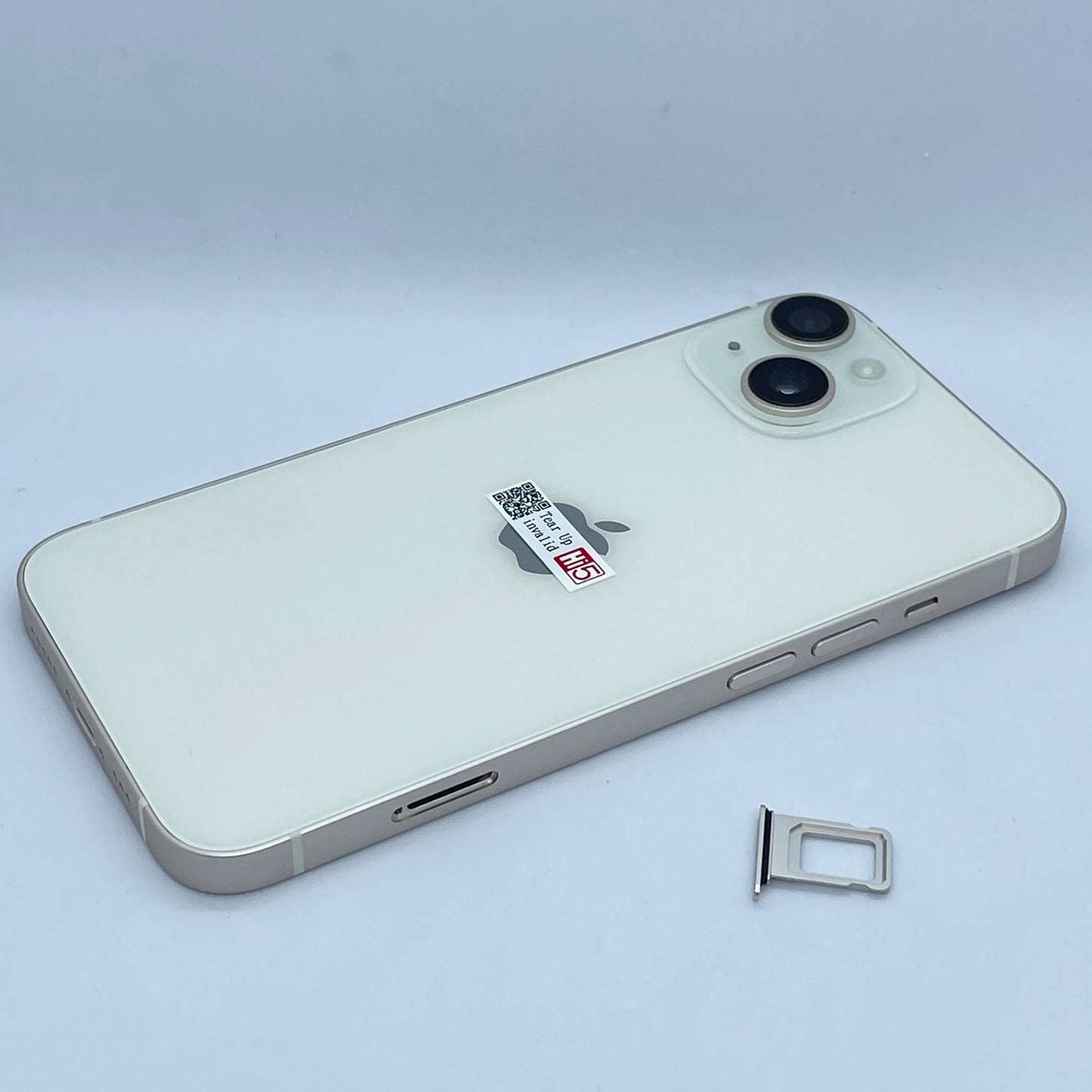 White smartphone with dual cameras on the back, laying on a flat surface. The SIM card tray is removed and placed next to the phone. Apple logo and a small sticker are visible.