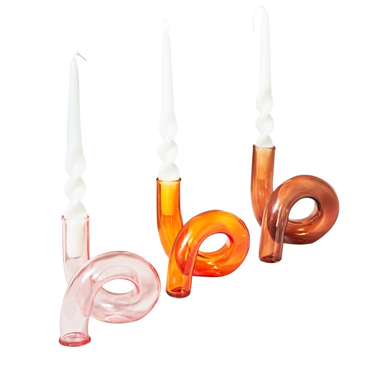 Three twisted white candles in curved, multicolored glass holders: pink, orange, and amber. The holders have a looped, modern design against a white background.