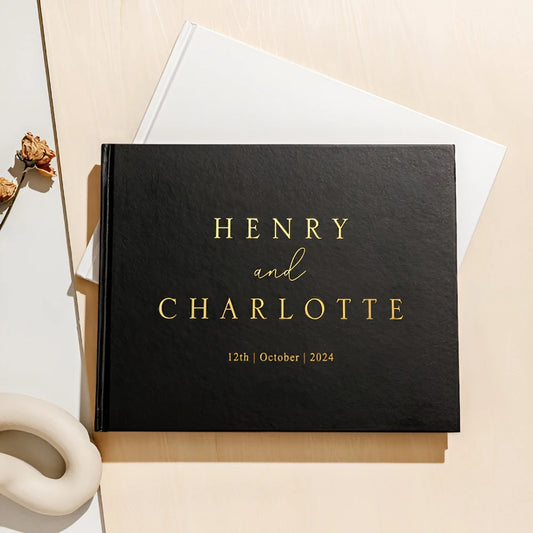 Black wedding album with "Henry and Charlotte" in gold letters, dated October 12, 2024. It's placed on a beige surface with dried flowers and a white ceramic piece nearby.