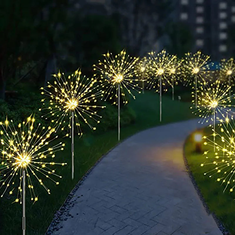 Pathway lined with glowing starburst lights resembling fireworks. The lights illuminate the winding path, surrounded by greenery, against a dim evening backdrop.