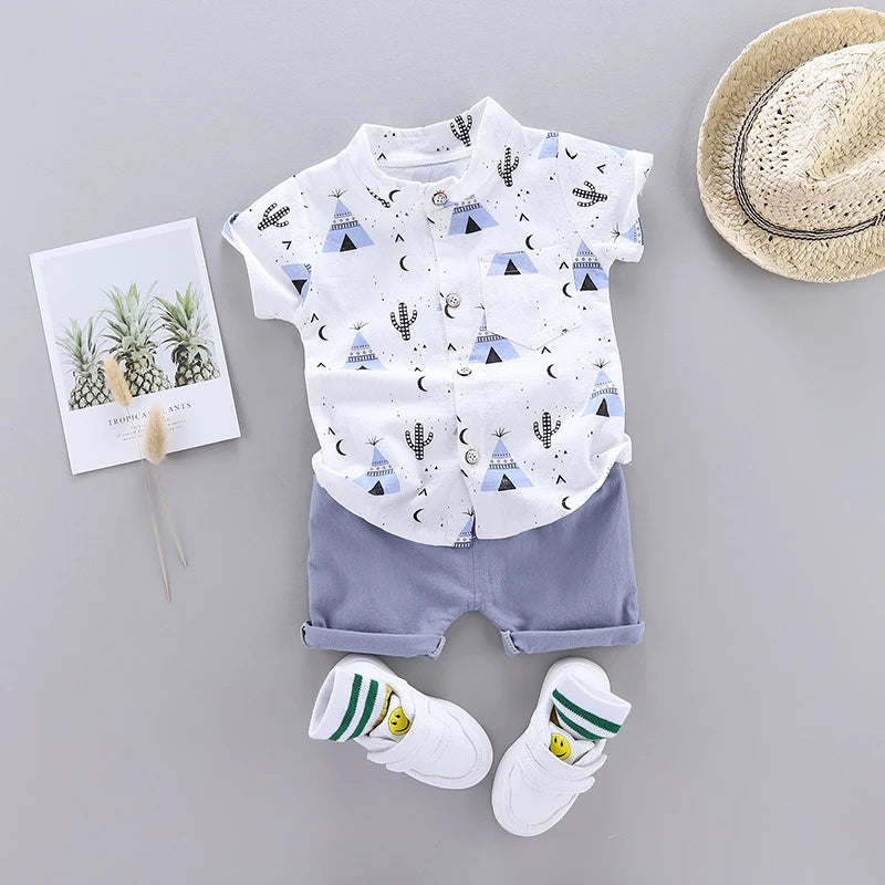 Children's outfit with a white shirt featuring blue teepees, cacti, and moons, paired with gray shorts. White sneakers with smiley socks. Includes a postcard with plants and a straw hat.