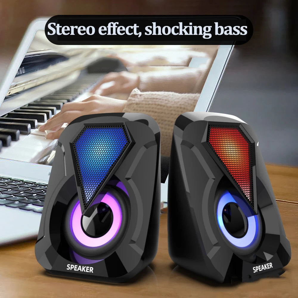 Two black desktop speakers with colorful LED lights sit on a wooden surface, in front of a laptop showing hands playing a piano, emphasizing stereo effect and powerful bass.