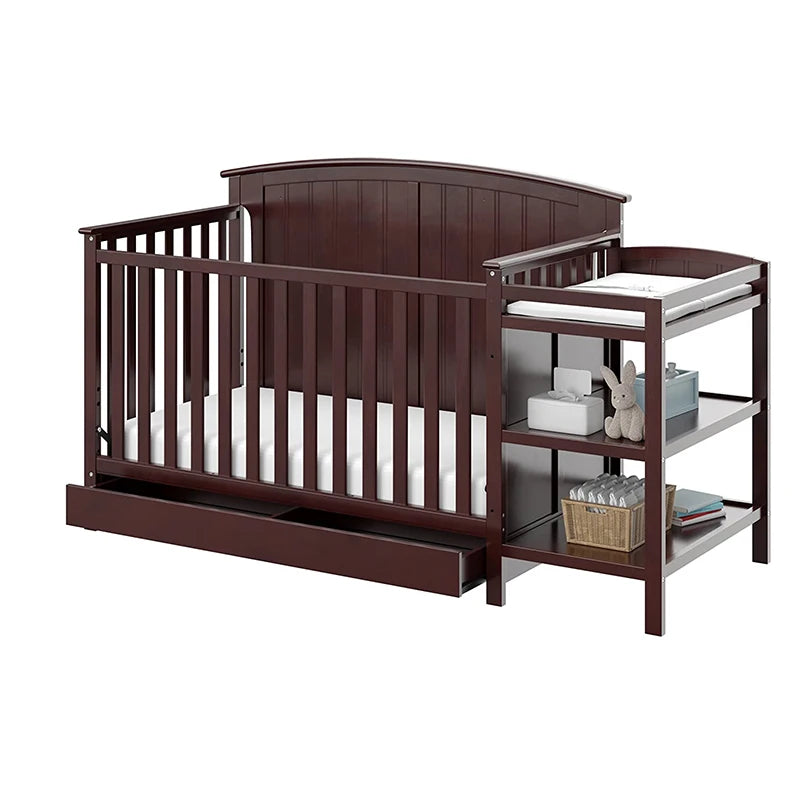 Dark brown convertible crib with slatted sides, an under-bed drawer, and an attached changing table with shelves. Accessories include a stuffed toy, diapers, and a decorative basket.