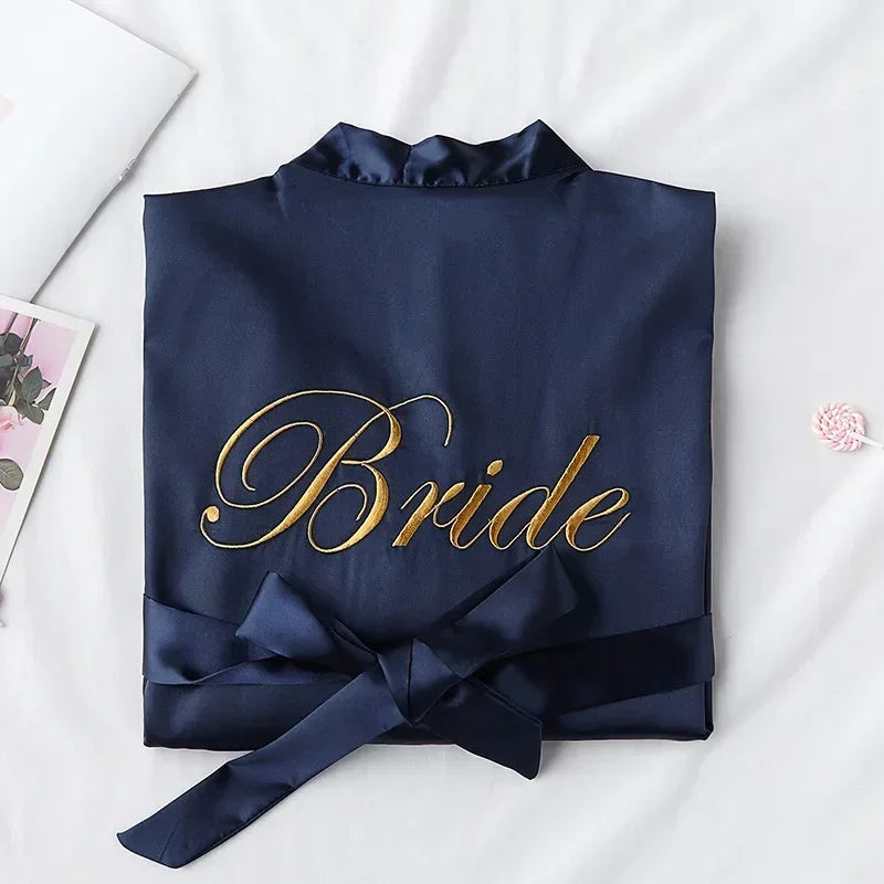 Navy satin robe with "Bride" embroidered in gold script, neatly folded with a tied sash. Background features a white surface, a pink floral card, and a small pink item.