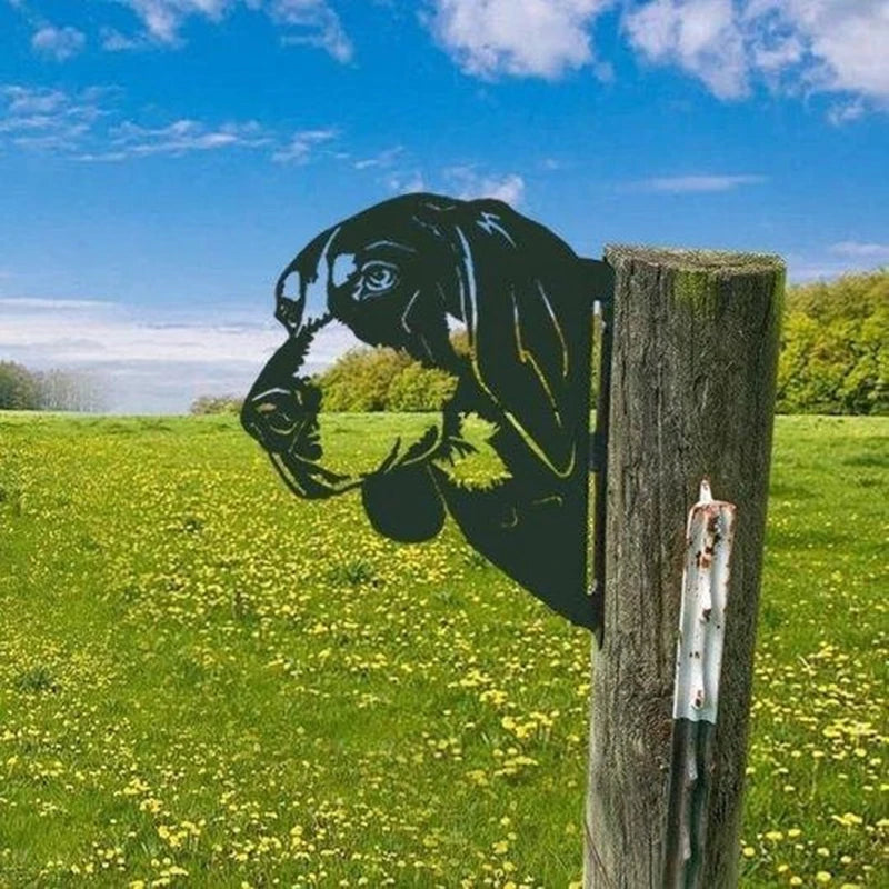 Metal silhouette of a dog's head mounted on a wooden post against a lush green meadow with yellow flowers and a blue sky with clouds.
