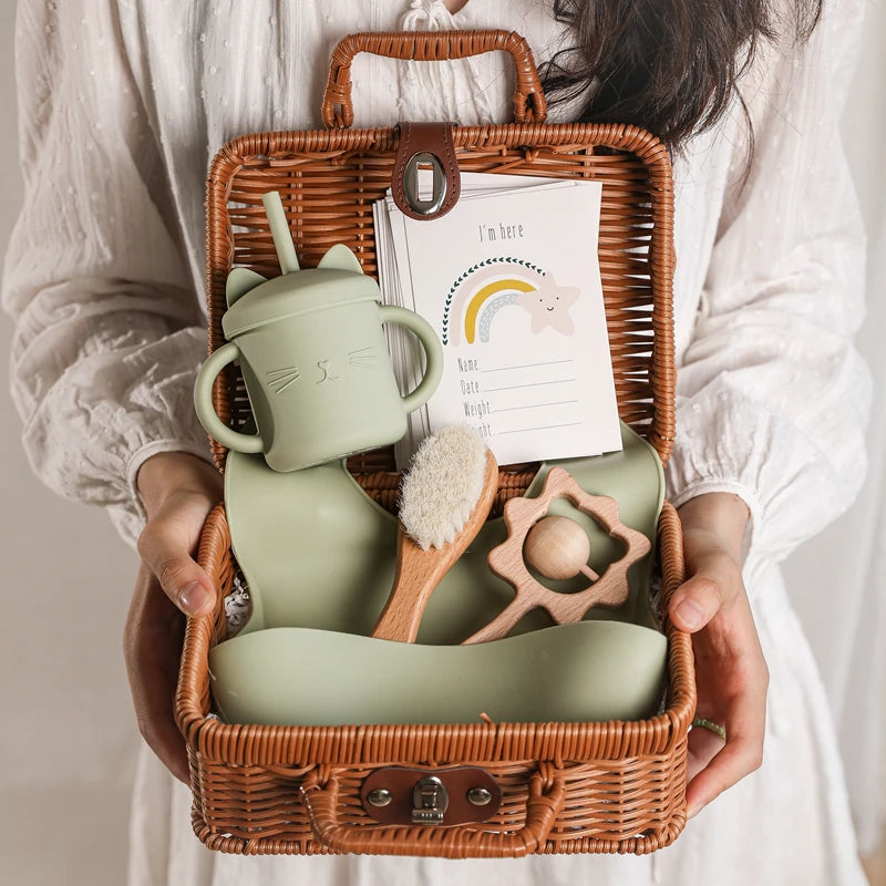 A person holds an open wicker basket containing a green sippy cup with cat ears, a baby brush, a teething toy, a silicone bib, and a birth announcement card with a rainbow design.