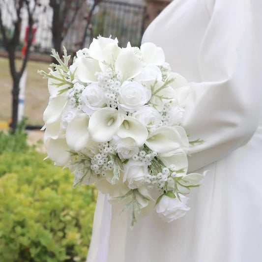 A bride holds a bouquet of white roses, calla lilies, and baby's breath. She wears a white gown, standing outdoors against a blurred background of greenery and trees.