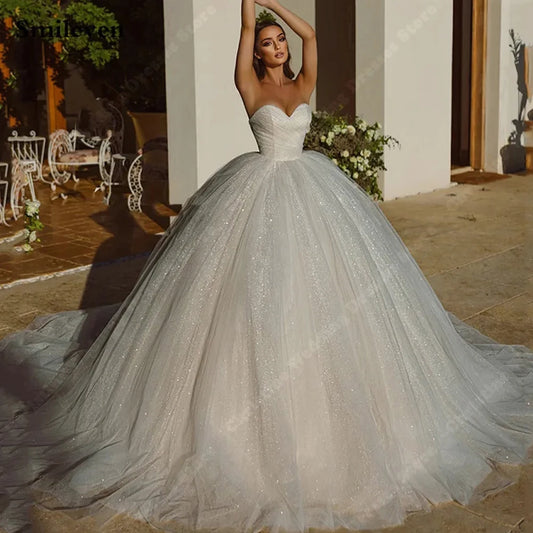 A woman in a strapless, shimmering white ball gown poses outdoors on a sunlit patio. The dress features a voluminous skirt, and the background includes elegant white furniture and greenery.