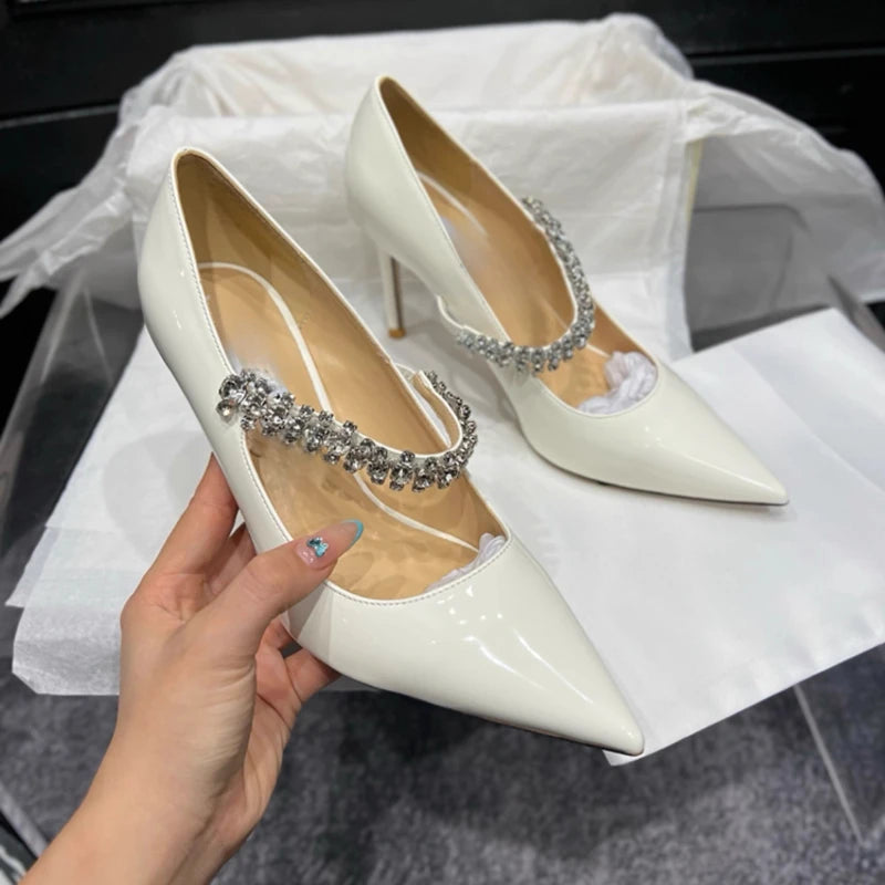 White pointed-toe high heels adorned with a sparkling rhinestone strap. One shoe is held in a hand with colorful nail art. Shoes are displayed on a white fabric.
