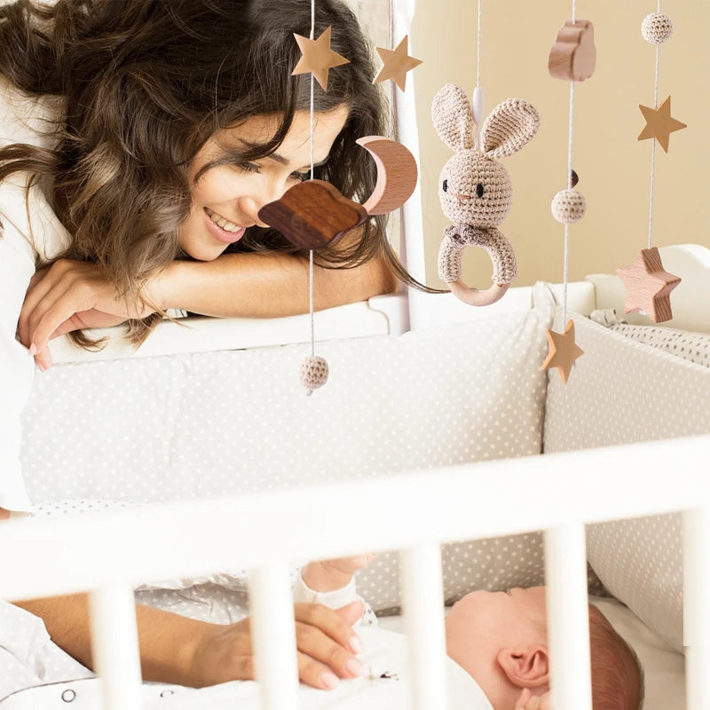 A woman smiles at a baby in a crib adorned with star and bunny-themed hanging toys. The setting is cozy with muted tones, creating a warm, nurturing atmosphere.