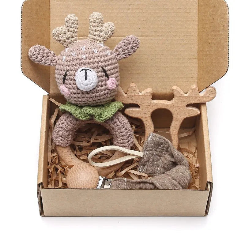 Crocheted deer toy with antlers and green collar, wooden teether, and fabric pacifier clip in a cardboard box with shredded paper packaging.