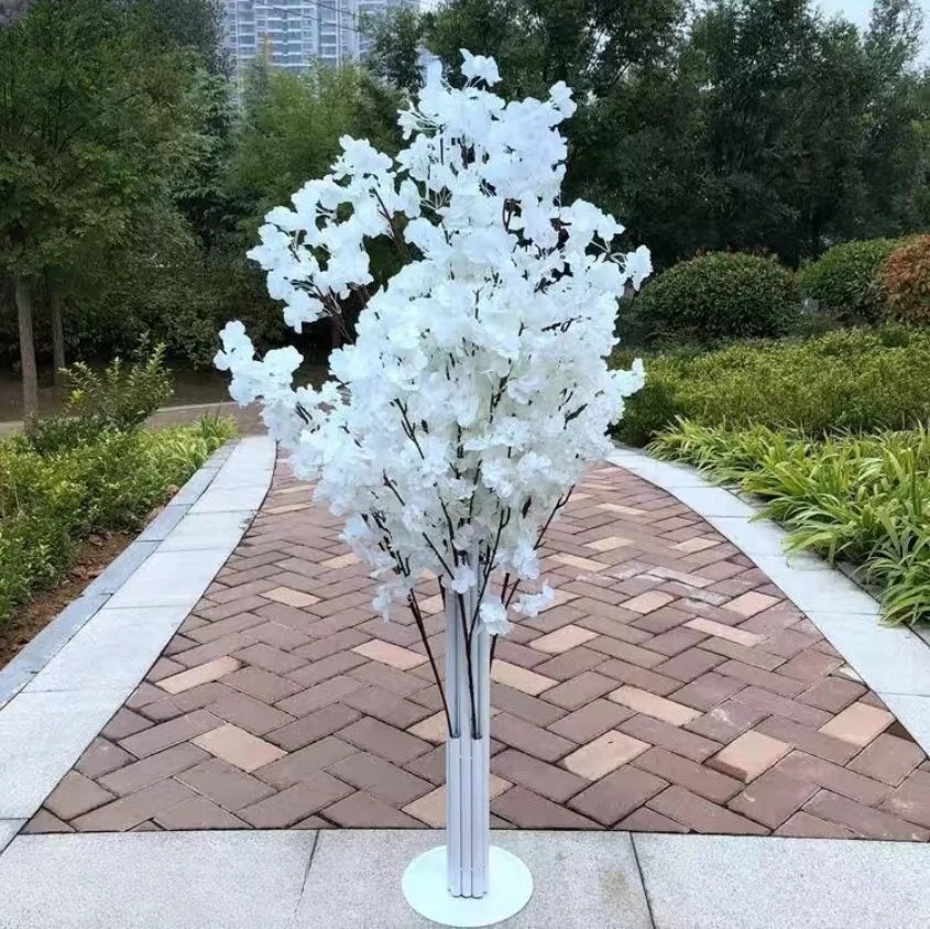 A small white artificial tree with numerous branches, set in a modern pot, stands on a brick pathway in a lush garden with greenery and trees in the background.