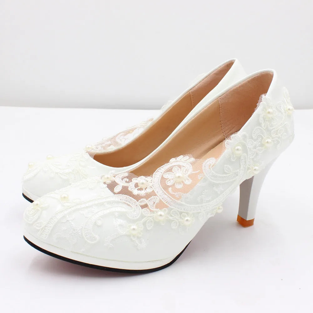 Elegant white high-heeled shoes with intricate lace detailing and small pearl accents on the sides, set against a plain white background. Perfect for formal occasions.