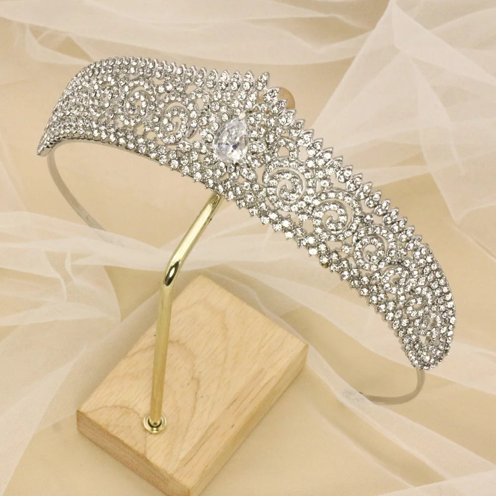 Ornate silver tiara adorned with intricate swirling patterns and sparkling rhinestones, displayed on a wooden stand with a soft fabric backdrop.