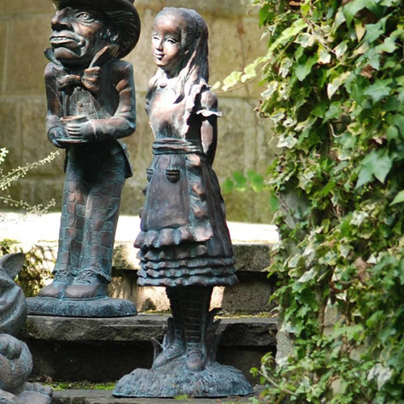 Bronze statues of a man and a girl dressed in old-fashioned clothing stand on stone steps surrounded by ivy. The man wears a hat and bow tie, while the girl has a braided hairstyle.