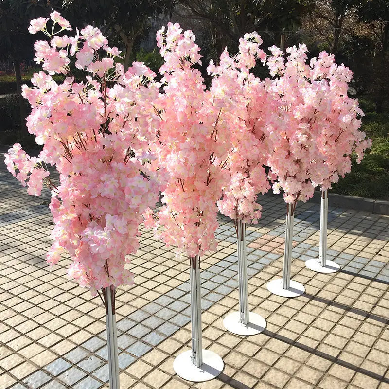 Four artificial cherry blossom trees with light pink flowers in full bloom, arranged in a line on a tiled outdoor surface, with green foliage in the background.
