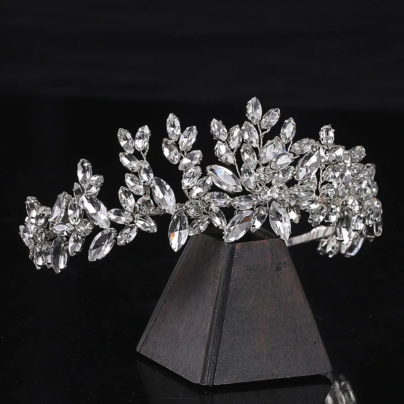 Elegant silver tiara adorned with sparkling crystals, arranged in a floral pattern, displayed on a dark wooden stand against a black background.