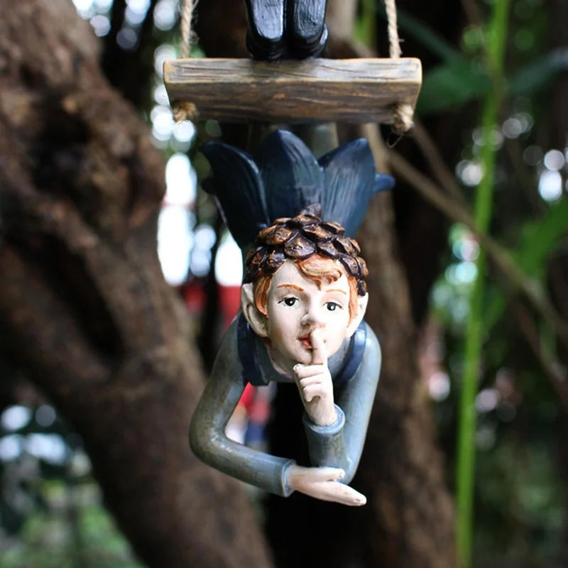 Hanging garden gnome figurine with a finger over its lips, signaling for silence. It wears a dark outfit and appears upside down against a blurred, leafy tree background.