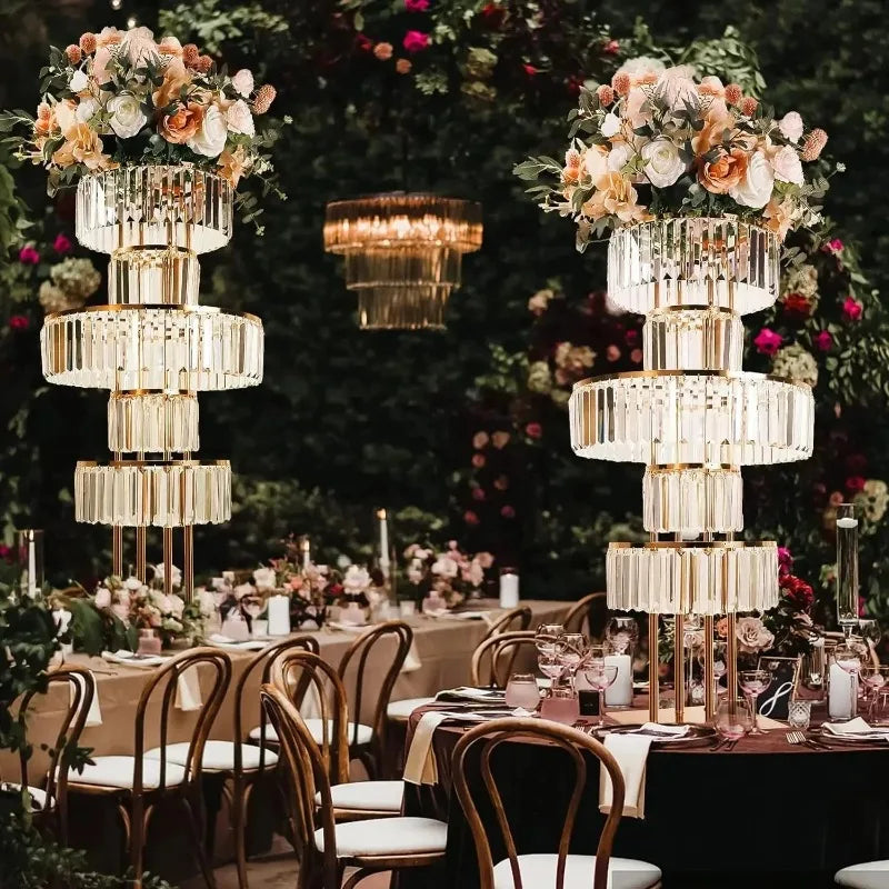 Elegant wedding reception table with crystal chandeliers adorned with flowers. Round tables are set with glassware and surrounded by wooden chairs, against a lush floral backdrop.